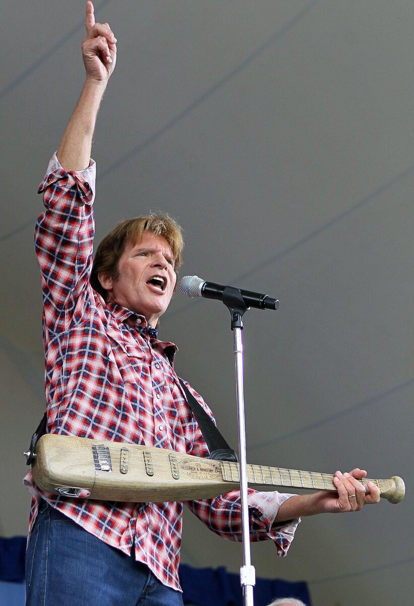 John Fogerty with his arm raised triumphantly, bat-shaped guitar in hand, on an outdoor stage
