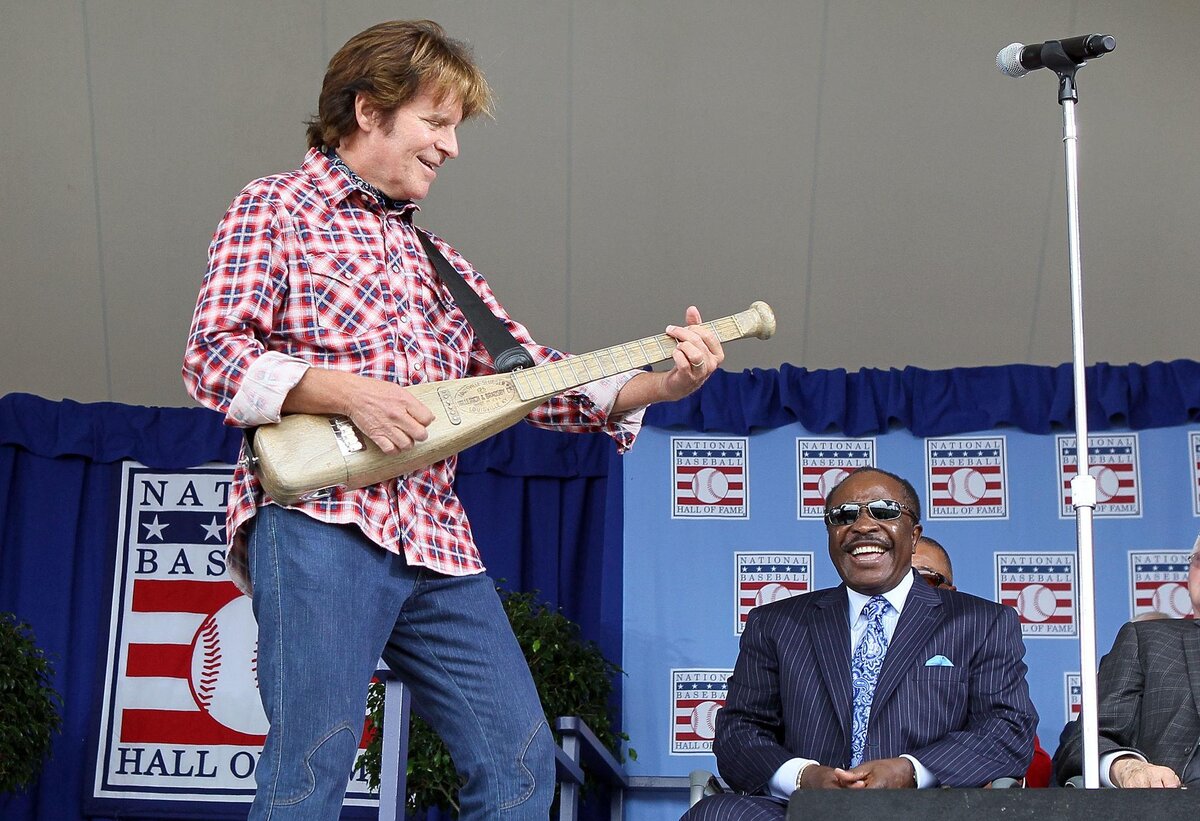 John Fogerty playing his baseball-bat guitar beside the National Baseball Hall of Fame logo