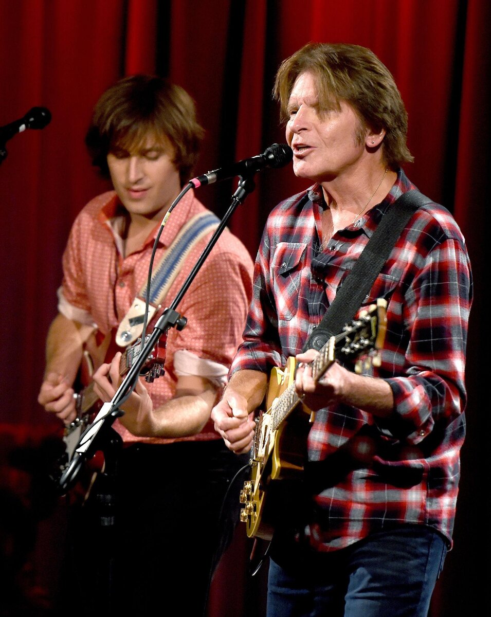 John Fogerty in a plaid shirt playing guitar on stage beside his son