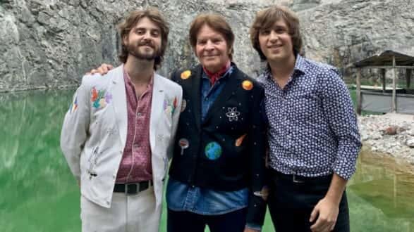 John Fogerty flanked by his sons Shane and Tyler outdoors by green water