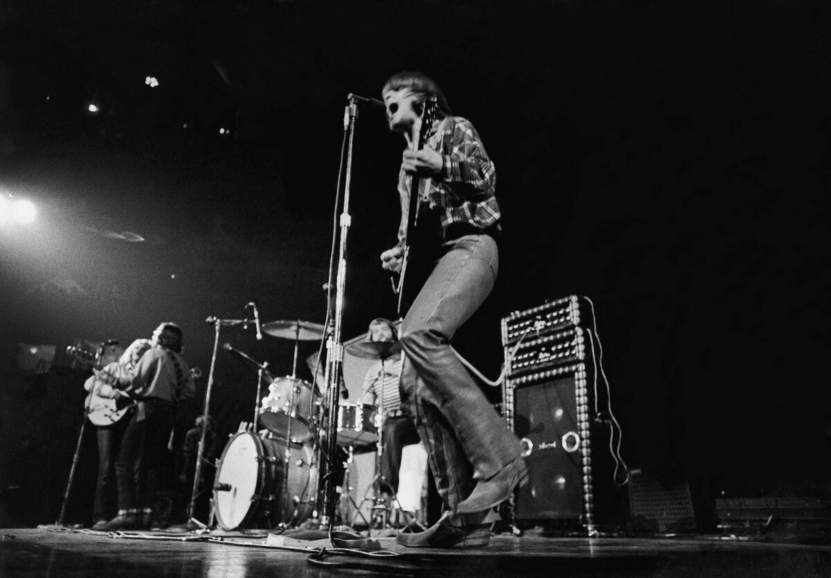 John Fogerty mid-performance with Creedence Clearwater Revival, black-and-white stage shot with drums and amplifiers behind him