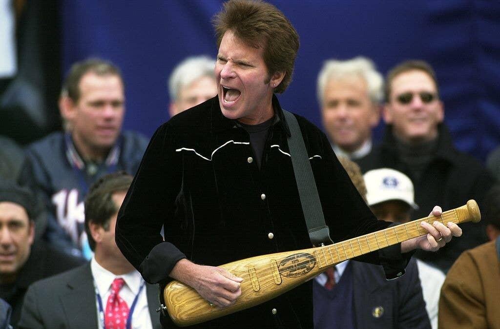 John Fogerty on stage with a baseball-bat-shaped guitar at a baseball celebration, surrounded by onlookers